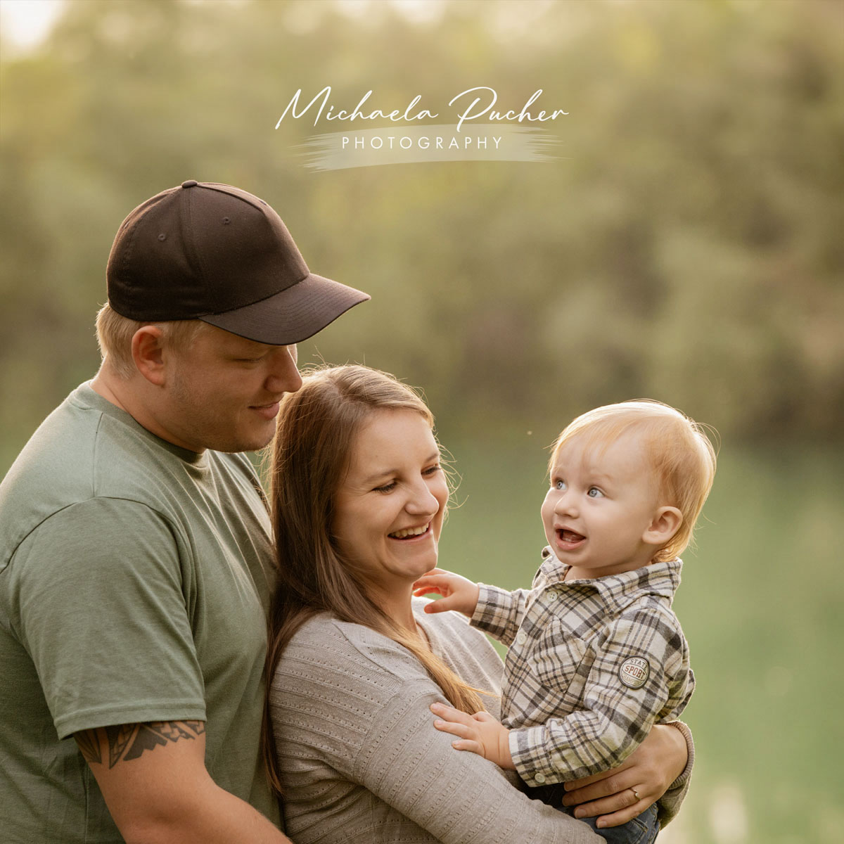 Mutter hält ihren kleinen Sohn im Arm, während der Vater liebevoll danebensteht; das Kind lacht die Mutter begeistert an, umgeben von weichem Abendlicht am See.